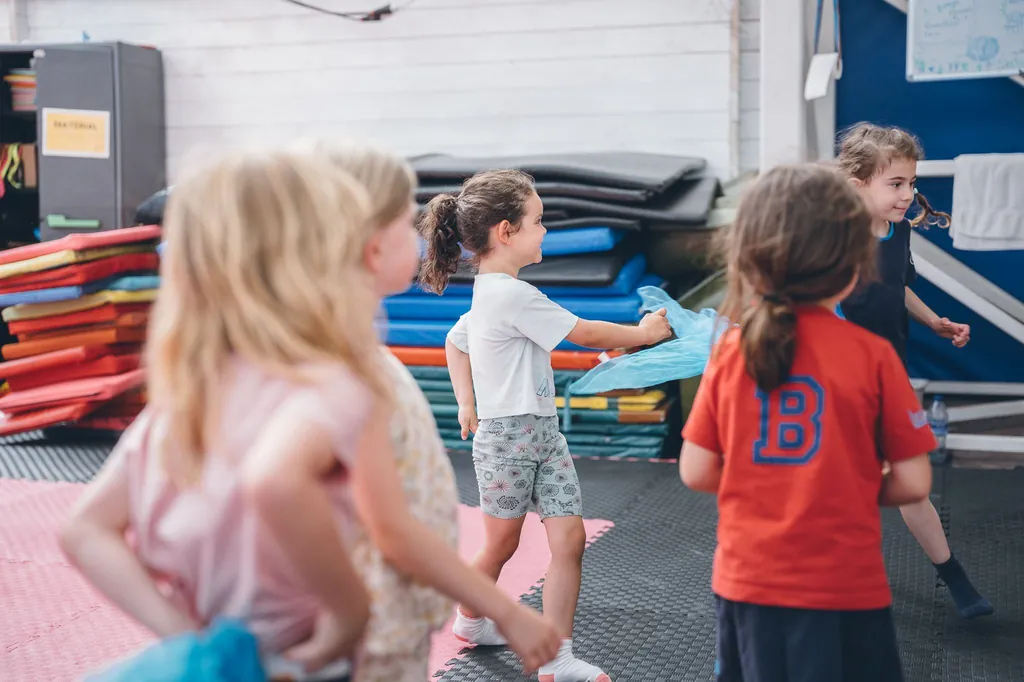 Varias niñas y niños juegan con pañuelos de colores sobre el tatami rosa durante una clase de circo.